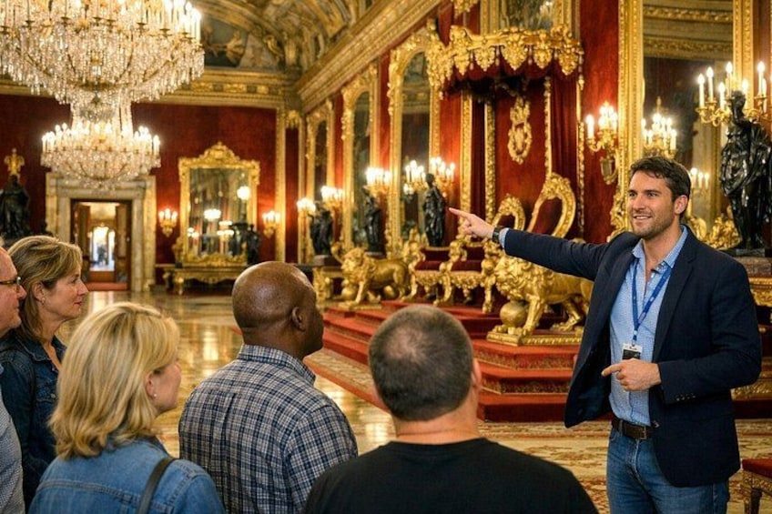 Een man wijst naar een gouden troon in een weelderige ruimte met kroonluchters en versierde decoraties. 1
