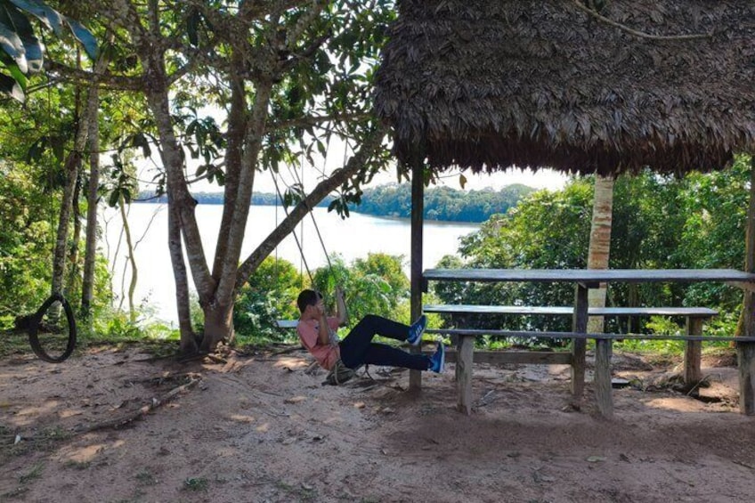 A person relaxing on a bench under a thatched roof, a hammock, and a view of a lake. 4