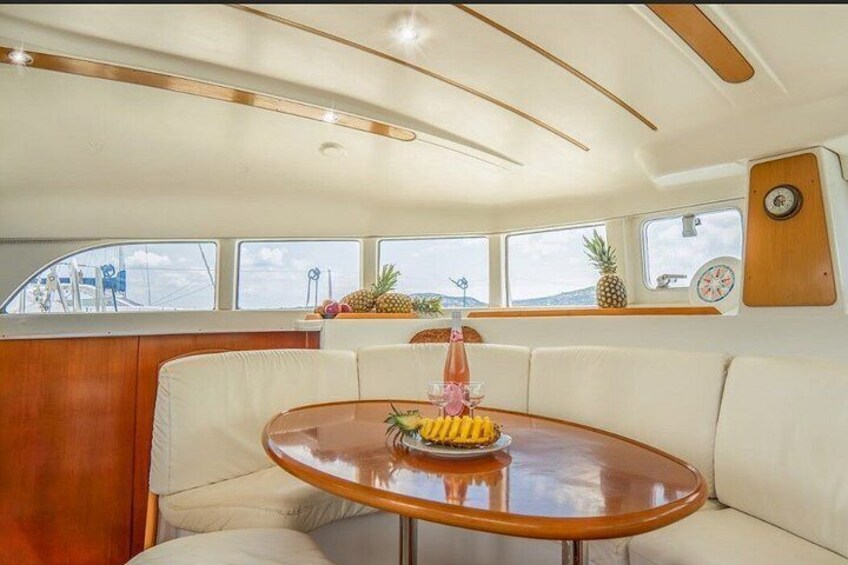 A boat interior with a round wooden table, white cushioned seating, and windows with tropical decor. 4
