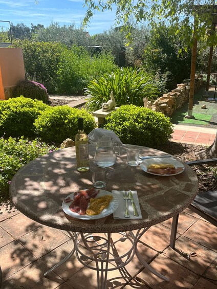 A round table with plates of food, glasses, and a bottle in a garden setting. 2