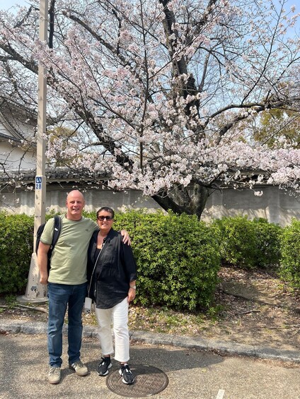 Two people standing in front of a cherry blossom tree with a building in the background. 2
