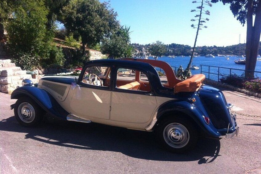 En klassisk bil parkerad vid en strandpromenad med fri utsikt över havet och båtarna. 3