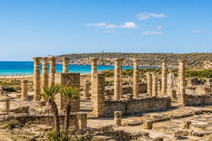 Ancient ruins with columns and a statue, located near a beach with clear blue waters. 2