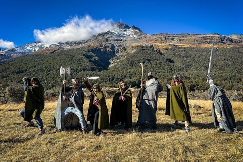 Un gruppo di persone in costumi medievali con spade e scudi in un prato erboso, con montagne sullo sfondo. 1