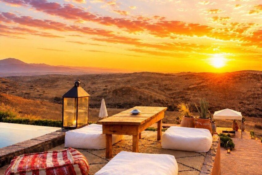 A sunset over a desert landscape with a wooden table and chairs, a lantern, and potted plants. 1