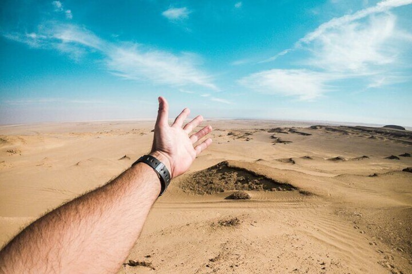 A hand reaching out towards a desert landscape with sand dunes under a clear sky. 3