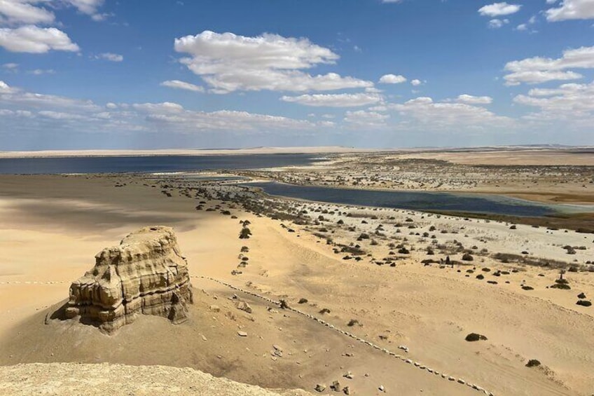 A desert landscape with a prominent rock formation, scattered boulders, and a distant shoreline. 2