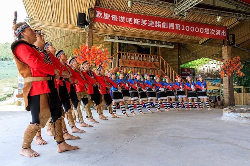A group of people in traditional attire performing a dance on a stage with a backdrop of text and a wooden structure. 4