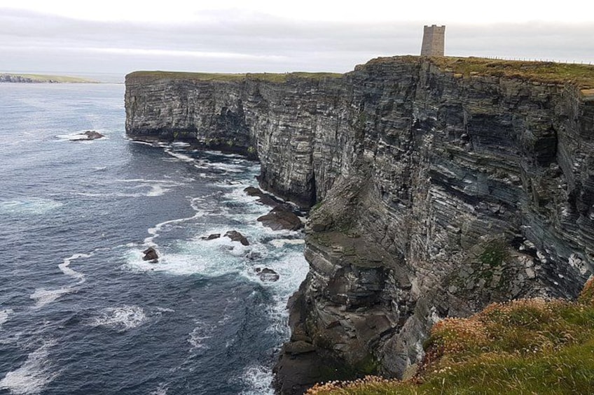Un acantilado costero con una torre en la cima, olas rompiendo en la base y una zona cubierta de hierba en el borde. 1