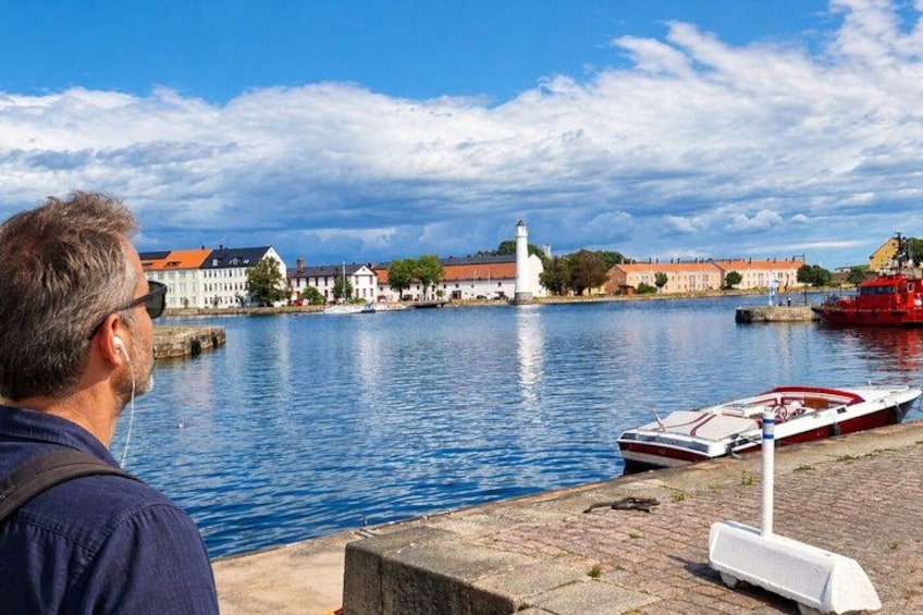 Un hombre observa un río tranquilo con barcos, edificios y un faro. 4