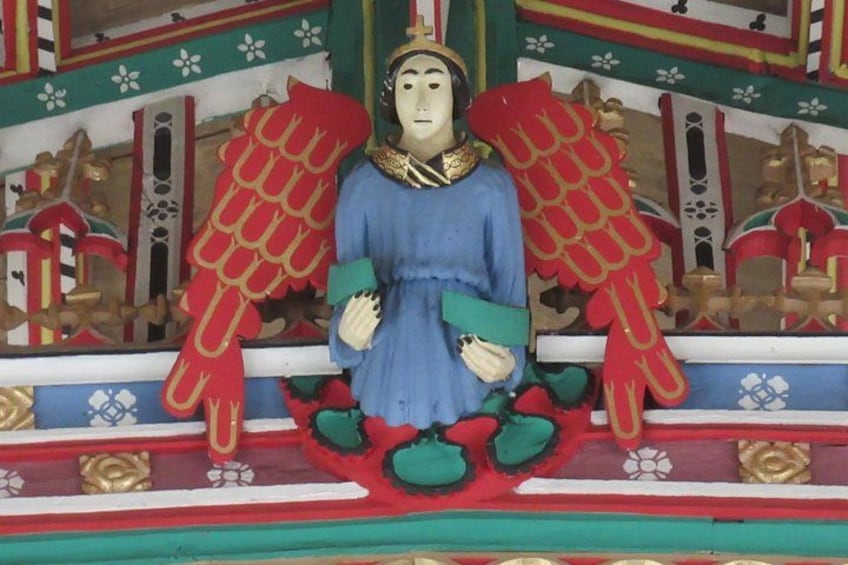 En statue af en engel med røde vinger og en blå kappe, siddende på en platform med detaljeret udsmykning. 1