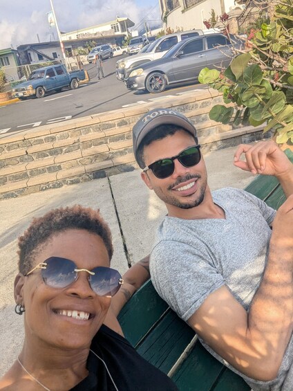 Two people sitting on a bench with a street and parked cars in the background. 2