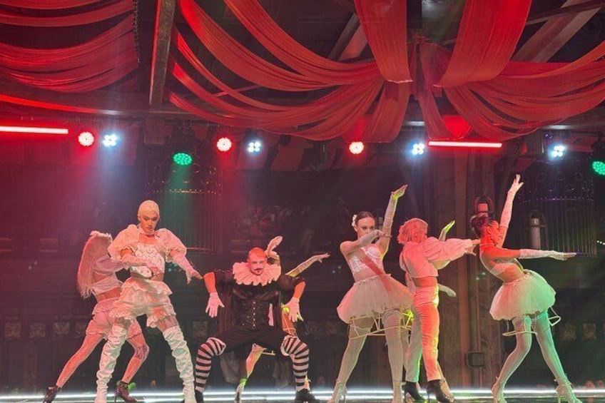 Un gruppo di ballerini in costumi elaborati si esibisce su un palcoscenico con tendaggi rossi e luci da palcoscenico. 2