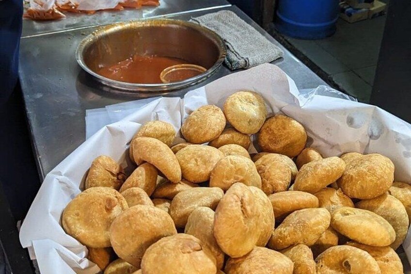 Een mand met goudbruine gefrituurde deegballen, een metalen kom met saus, en een blauwe vat op de achtergrond. 2