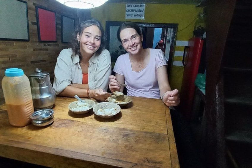 Twee personen zitten aan een houten tafel met bakjes eten en een metalen pan. 3