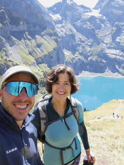 A person with a backpack and another person in a cap, standing on a grassy hill with a mountainous landscape and a lake in the background. 5