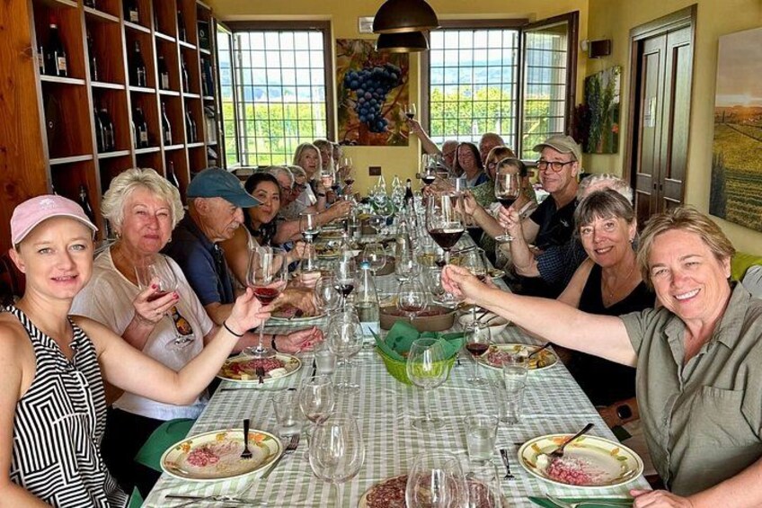 A group of people seated at a long table with wine glasses and plates, in a room with wooden shelves and a painting on the wall. 3