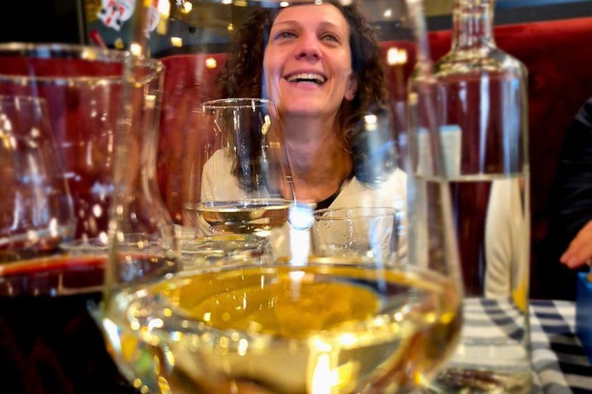 A person seated at a table with a large, clear glass bowl filled with a golden liquid, possibly wine, and several bottles in the background. 3