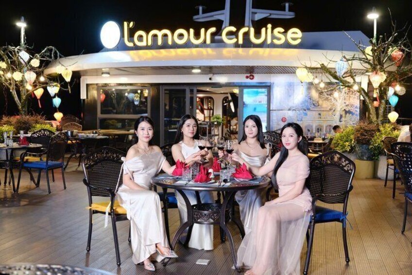 Un espace extérieur aménagé pour les repas, avec des tables et des chaises, et au fond, on aperçoit un restaurant appelé « L'amour cruise », le tout agrémenté de guirlandes lumineuses. 3