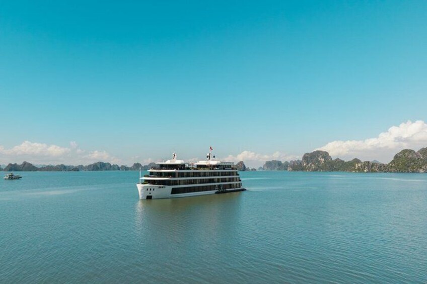 Un grand navire de croisière blanc navigue sur des eaux calmes, sous un ciel dégagé, avec des reliefs terrestres visibles au loin. 1