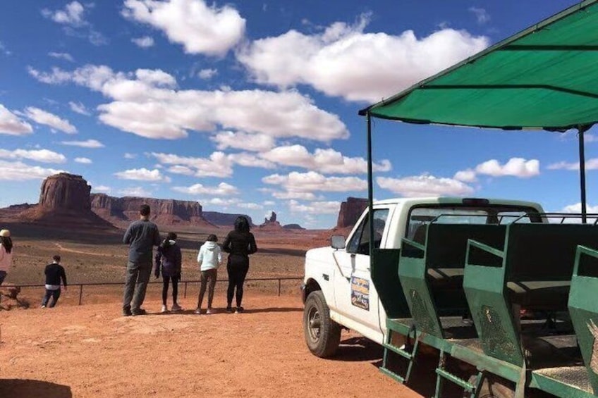 Un groupe de personnes se tient près d’un camion blanc à benne, dont les sièges sont verts, dans un paysage désertique caractérisé par des formations rocheuses rouges et un ciel dégagé. 2