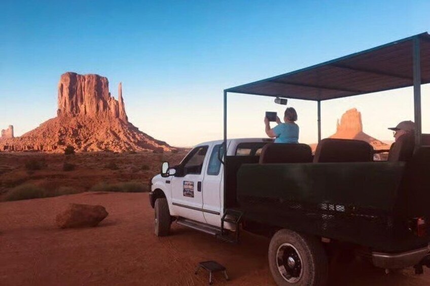 Un camionnette blanche à plateau, dotée d’une cabine de toit, est garée dans un désert, avec des formations rocheuses en arrière-plan. 1