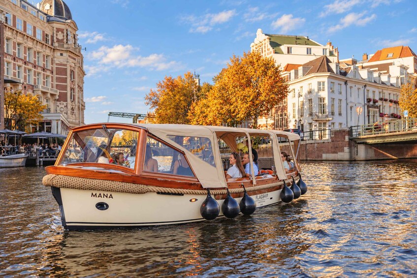 A boat named 'MANA' on a river with buildings and autumn trees in the background. 4