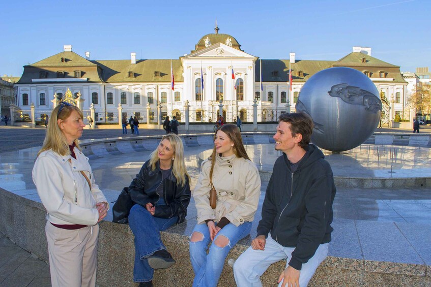 Four people sitting on a bench in a plaza with a large sculpture and a historic building in the background. 5