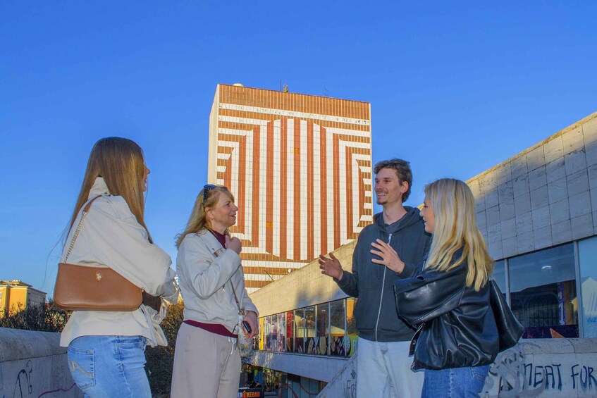 Four people standing outdoors with a large building in the background. 2