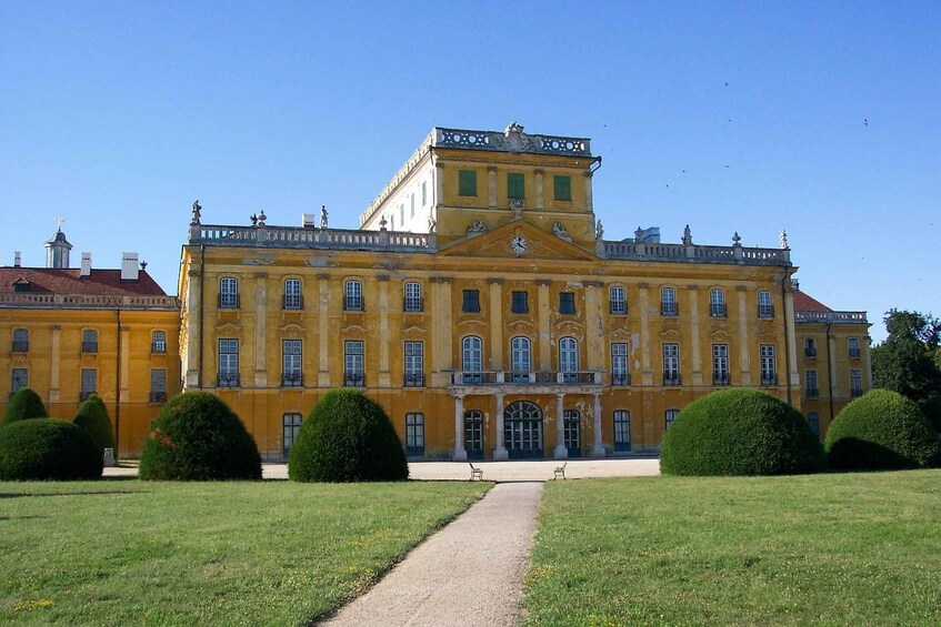 A large yellow building with a central entrance and symmetrical windows, surrounded by well-manicured lawns and hedges. 3