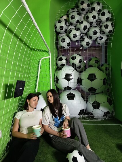 Two people sitting on artificial turf with a soccer goal and multiple soccer balls in the background. 3