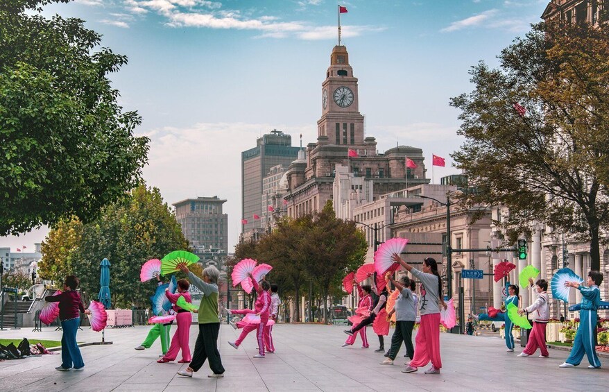 A group of people performing a traditional dance with fans in an urban square with historic buildings in the background. 2