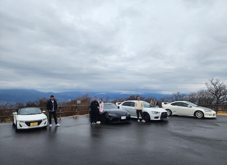 A group of people standing beside four cars in a parking lot with a mountainous backdrop. 5