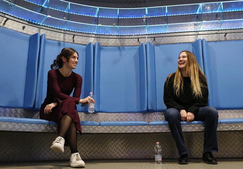 Two people sitting on blue benches in a room with a patterned floor and a curved ceiling with lights. 2