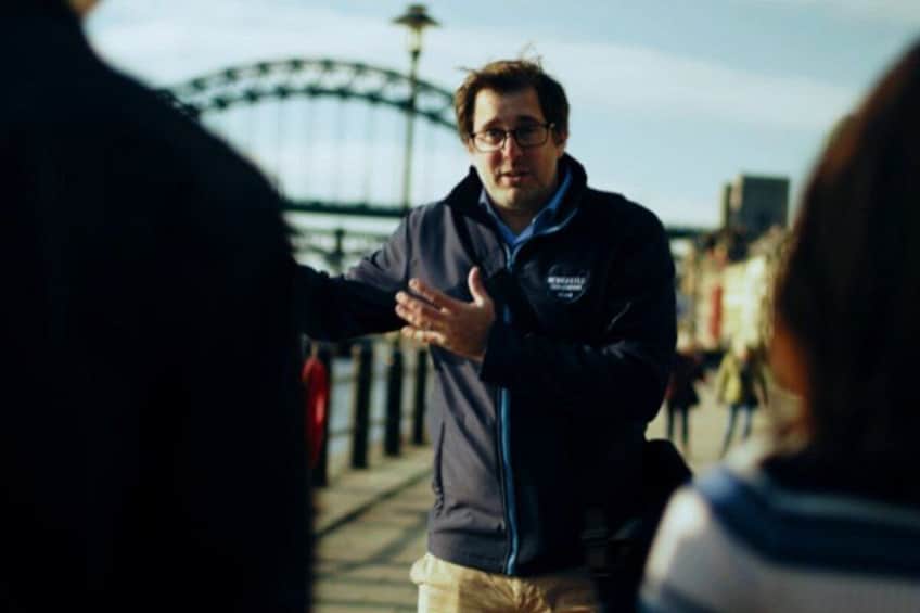 A person gesturing with hands, wearing a jacket with a logo, standing on a bridge with a clear sky in the background. 2