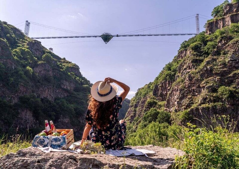 A person sitting on a rock with a picnic setup, overlooking a gorge and a bridge. 5