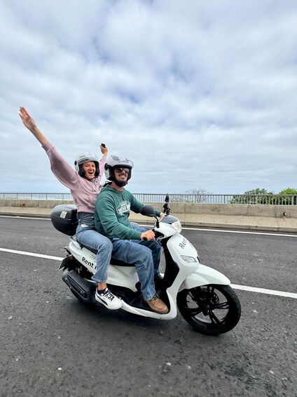 Two people on a white scooter with 'Rent' written on it, riding on a road with a metal fence and trees in the background. 1