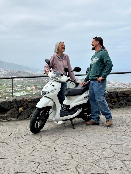 Two people standing beside a white scooter with 'rent me' and 'TVR' on it, overlooking a cityscape with buildings and hills. 4
