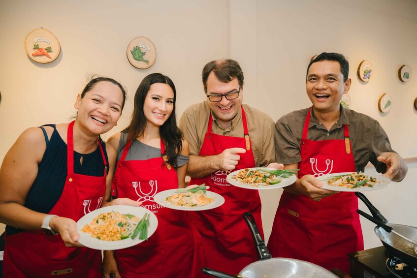 Four people in red aprons holding plates of food, with kitchen utensils and decorative plates on the wall. 1