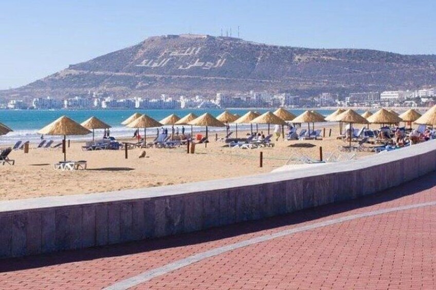 A beach with sun loungers and thatched umbrellas, a concrete walkway, and a hill in the background. 1