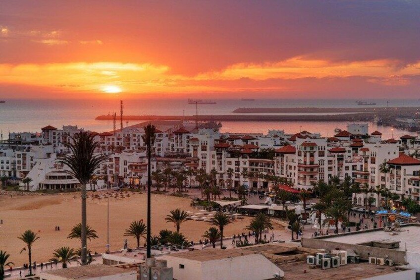 A coastal cityscape at sunset with buildings, palm trees, and a beach. 2