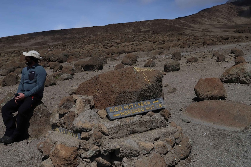 A person sitting on rocks with a sign indicating distances to Kibo Hut and Horomoe Hut. 2