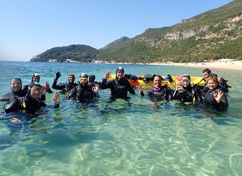 A group of people in scuba diving gear posing in shallow water with a boat and a mountainous coastline in the background. 4