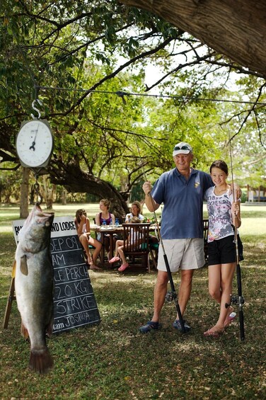 A man and a woman fishing under a tree with a fish on a hook, a weighing scale, and a sign indicating a fishing competition. 1