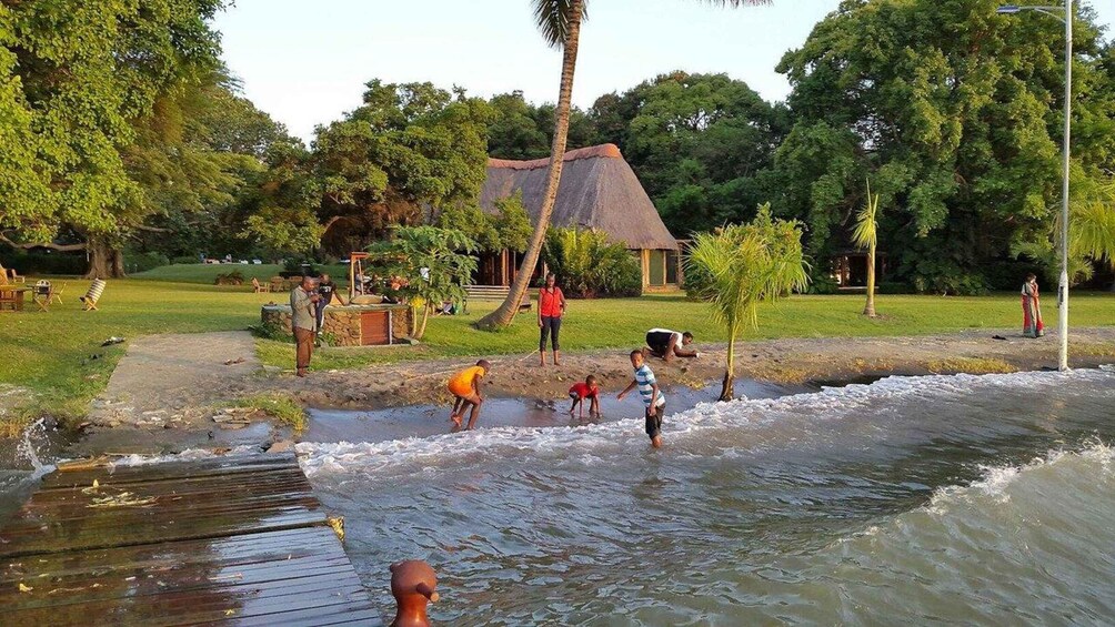 A river with people playing in the water, a thatched-roof hut, and lush greenery. 4