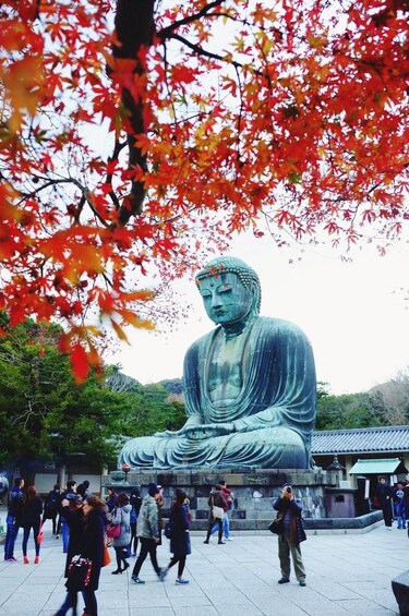 A large Buddha statue surrounded by people in a park with autumn leaves. 1
