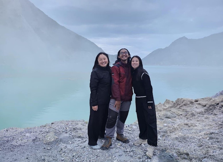 Three people standing on a rocky shore with a lake and mountains in the background. 4