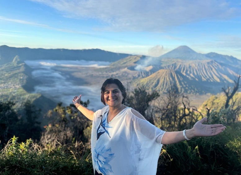 A person standing with arms outstretched, overlooking a volcanic landscape with visible smoke and distant mountains. 1