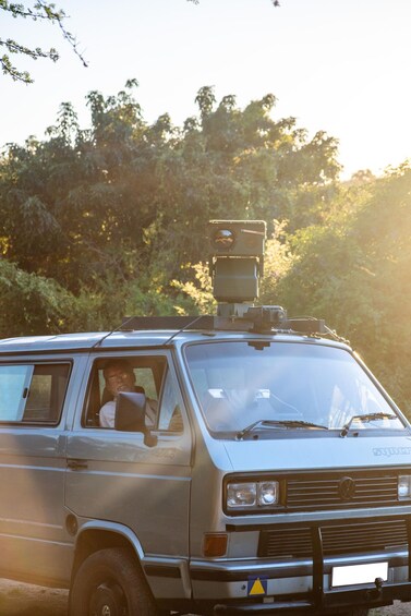 A silver van with a mounted camera on the roof, parked in a wooded area. 5