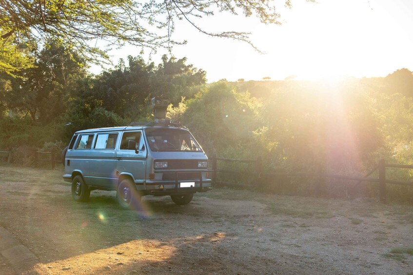A vintage van parked on a dirt path with trees and a wooden fence in the background. 2